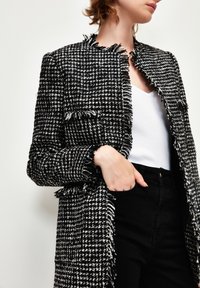 Černobílý tweedový blazer s třepenými okraji, otevřený vpředu, se dvěma kapsami, v kombinaci s bílým topem a černými džíny. Hladká textura.