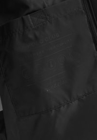 Närbild av svart Polo Club-jacka tyg som visar sydd etikett med ikoner för andningsförmåga, temperatur, vattenbeständighet och hållbarhet.