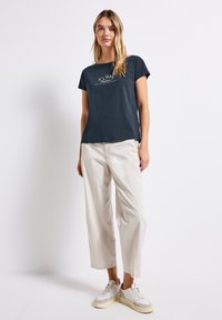 T-shirt navy à manches courtes avec un graphisme sur le devant, associé à un pantalon large beige clair et des baskets blanches avec des accents texturés.