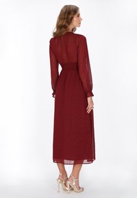 Rochie bordeaux, cu mânecă lungă, cu talie încrețită, dintr-o țesătură transparentă, cu puncte texturate. Purtați cu sandale aurii cu toc. Vedere din spate.