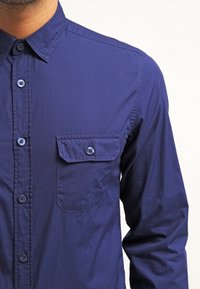 Marinblå långärmad skjorta i lätt tyg, med knäppning framtill, spetsig krage och en enda bröstficka.