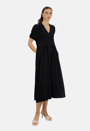 Kvinne står med hendene i lomma, iført en svart midi-kjole med knapper, puffarmer og hvite sandaler med hæl.