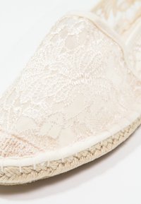 Espadrille crème à enfiler avec un superposé en dentelle, semelle en jute tissé et bout arrondi. Tissu texturé avec des motifs floraux et accents cousus.