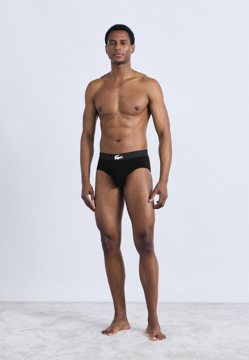 Μαύρο εσώρουχο τύπου σλιπ με γκρι ζώνη μέσης που έχει λευκό λογότυπο. Το μοντέλο στέκεται ξυπόλυτο σε ανοιχτό γκρι επιφάνεια.