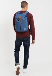Zaino in tessuto blu con parte superiore arrotondata, accenti in pelle, tasche frontali e spallacci regolabili, indossato sopra un maglione bordeaux e jeans scuri.