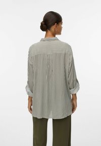Femme avec les cheveux en chignon portant une chemise longue à manches longues à rayures verticales et un pantalon vert foncé, vue de dos.