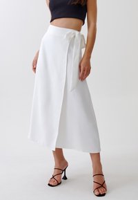 Jupe midi blanche avec un design enrobé, texture lisse et détail de nœud sur le côté. Associée à des talons noires à lanières, mettant en valeur une ouverture sur le devant.