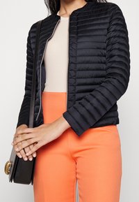 Black puffer jacket with horizontal quilted texture, zip closure, and round neckline. Paired with orange pants, carrying a black handbag.