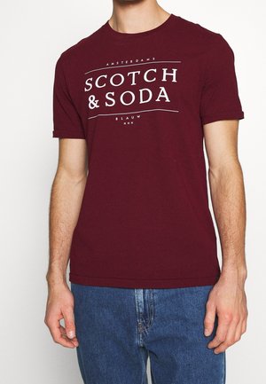 Uomo che indossa una T-shirt bordeaux a maniche corte con scritta bianca "Scotch & Soda" e jeans blu, in piedi davanti a uno sfondo semplice.