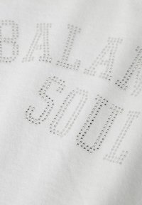 Biała bawełniana tkanina z napisem "BALANCE SOUL" wykonanego z srebrnych dżetów, prezentująca gładkie, teksturowane wykończenie.