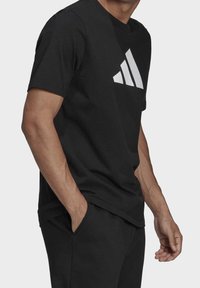 Muž v černém tričku Adidas s bílým logem, se založenýma rukama v kapsách, zobrazený od poloviny hrudníku po boky na jednobarevném pozadí.