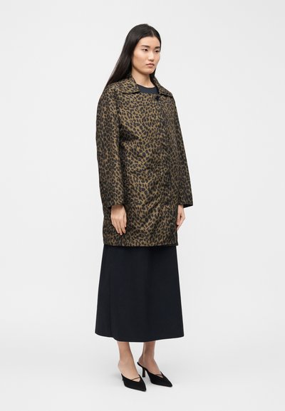 Γυναίκα που φοράει καφέ και μαύρο παλτό με λεοπάρ σχέδιο πάνω από μαύρη μακριά φούστα και μαύρες μυτερές γόβες, στέκεται μπροστά σε ένα απλό λευκό φόντο.