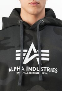 Primer plano de una persona que lleva una sudadera con capucha de camuflaje negro con el logo blanco "Alpha Industries Knoxville, Tennessee, EE. UU." y cordones de color gris oscuro.