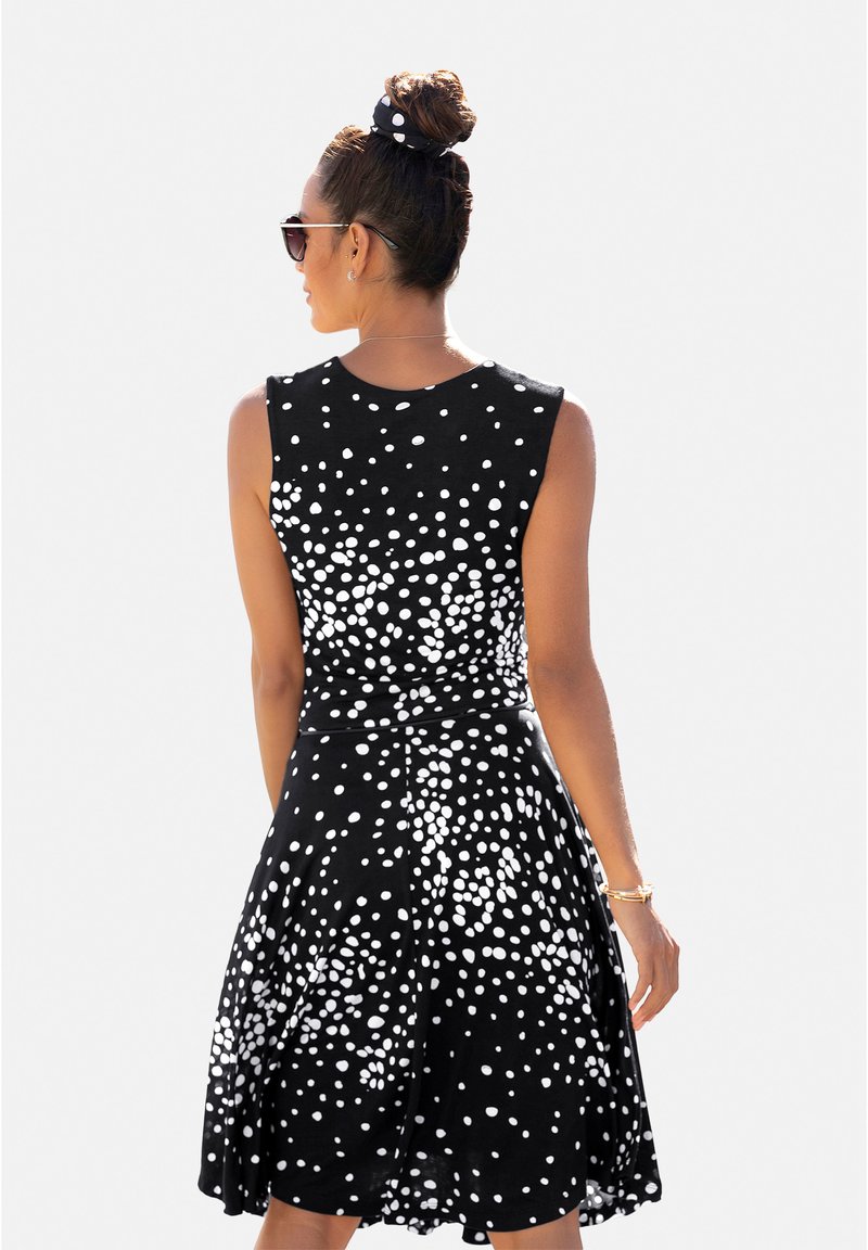 Schwarzes ärmelloses Kleid mit ausgestelltem Rock, das ein weißes Punkte-Muster aufweist. Kombiniert mit einem passenden Scrunchie und Sonnenbrille.