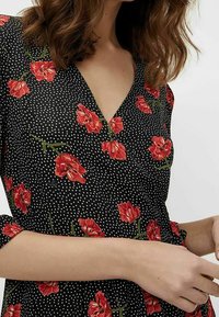 Robe noire à pois avec un motif floral rouge. Elle présente un décolleté en V et des manches courtes, fabriquée en tissu léger.