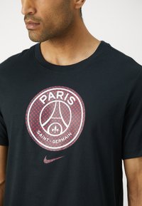 Sort bomulds t-shirt med et cirkulært Paris Saint-Germain-logo featuring et rødt geometrisk mønster. Inkluderer en lille Nike swoosh i rødt.