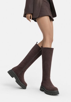 Frau trägt dunkelbraune kniehohe Stiefel mit dicken schwarzen Sohlen, kombiniert mit einem passenden Rock und Blazer.
