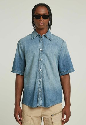 Mann mit geflochtenem Haar, der dunkle Sonnenbrillen, ein blaues Denim-Kurzarmhemd mit Knöpfen und beige Cargohosen vor einem einfarbigen Hintergrund trägt.