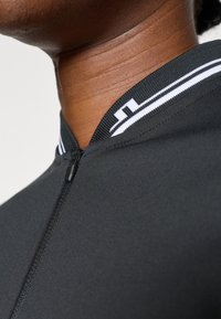 Melnā velosipēda krekls ar rāvējslēdzēju, kuram ir balts svītru apkakle un gluda, teksturēta audums. Tuvplāna skats atklāj detaļas.