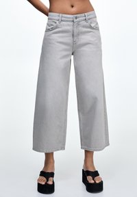 Personne portant un jean gris clair taille haute, jambes larges, coupé au-dessus des chevilles, avec des sandales noires à plateforme style tongs, sur un fond uni.