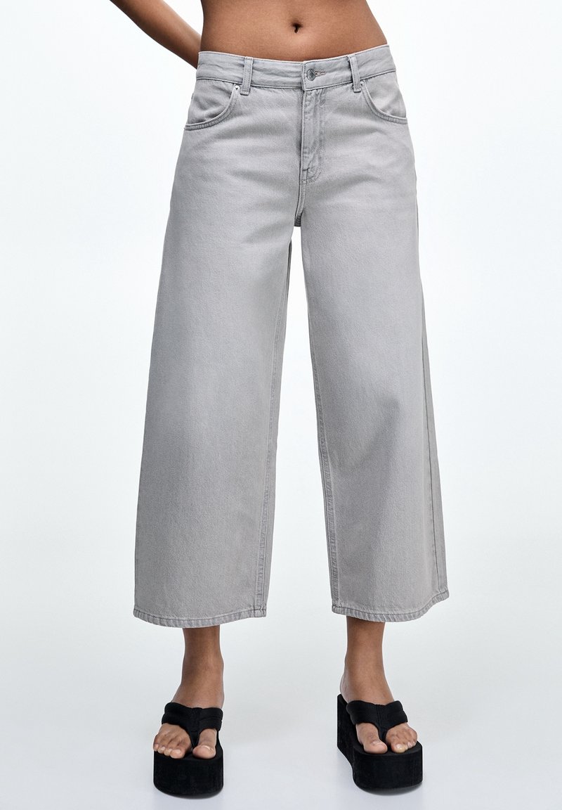 Personne portant un jean gris clair taille haute, jambes larges, coupé au-dessus des chevilles, avec des sandales noires à plateforme style tongs, sur un fond uni.