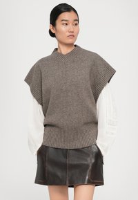 Junge Frau mit dunklen Haaren, die nach hinten gebunden sind, trägt einen grauen Strickwesten über einer weißen Bluse und einem dunkelbraunen Lederrock, die Hände in den Taschen.