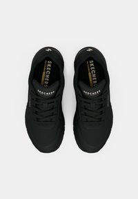 Schwarze Sportschuhe mit glatter Oberfläche, ausgestattet mit Schnürung, strukturierten Akzenten und einer gepolsterten Kragenpartie für Komfort. Logo auf der Zunge sichtbar.