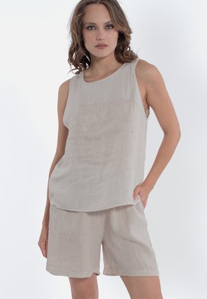 Donna con capelli castani fino alle spalle che indossa una maglia beige senza maniche larga e pantaloncini abbinati, in piedi con una mano in tasca.