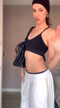 Jeune femme posant avec les lèvres pincées, portant un soutien-gorge de sport noir, un pantalon blanc, un bandeau noir, des boucles d’oreilles en anneaux dorés et tenant un sac noir.