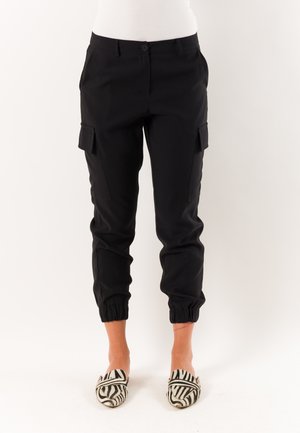 Pantaloni neri in stile cargo con polsini elasticizzati indossati con scarpe slip-on stampa zebra su una persona su sfondo neutro.