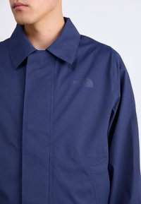 Vīrietis ar tumši zilu North Face jaku ar apkakli, redzama augšējā krūtis un daļa sejas pret vienkāršu fonu.