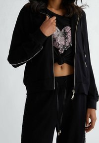Zwarte ritssluiting hoodie met glanzende zwarte accenten, gelaagd over een zwart T-shirt met een roze, textuur hartontwerp. Gestyled met zwarte joggers.