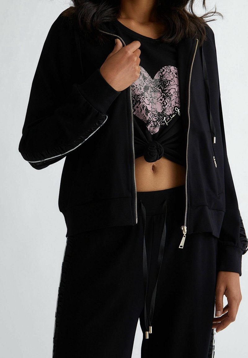 Zwarte ritssluiting hoodie met glanzende zwarte accenten, gelaagd over een zwart T-shirt met een roze, textuur hartontwerp. Gestyled met zwarte joggers.