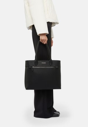 Personne portant un pantalon noir et une veste blanche, tenant un grand sac fourre-tout en cuir noir avec une poche zippée et des poignées doubles.
