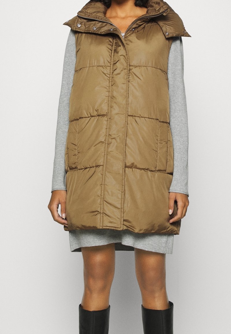 Veste matelassée marron avec col montant, zip à l'avant et deux poches latérales. Portée par-dessus une robe à manches longues grise, complétée par de hautes bottes noires.