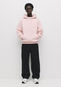 Helles rosa Kapuzenshirt mit einer Kängurutasche, kombiniert mit lockeren schwarzen Jogginghosen und reflektierenden Turnschuhen. Schlichte Hintergrund.