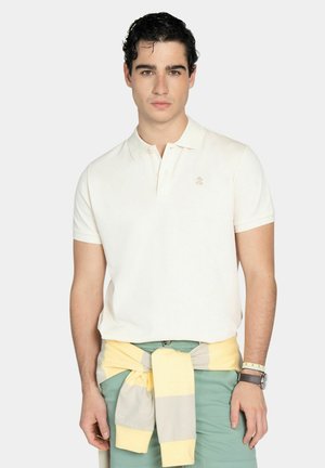 Joven con cabello oscuro, vestido con una camiseta polo blanca y pantalones verdes, suéter amarillo-gris atado alrededor de la cintura, de pie frente a un fondo liso.