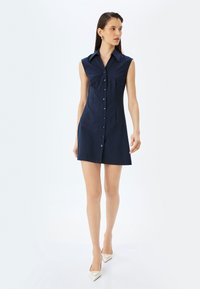 Merennavynen sleeveless-mekko, jossa on kauluksellinen pääntie ja napitus edessä, omaa kapean muotoilun ja suoran helman. Sujuva kankaan rakenne.
