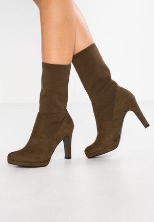 Botas de tobillo de ante marrón con un upper de tejido ajustado tipo calcetín, punta afilada y un alto tacón de aguja. Textura suave con un sutil detalle de costura.