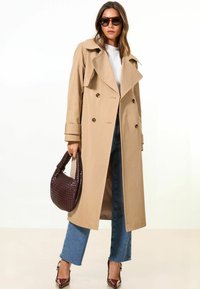 Trench beige con botones de doble botonadura, amplios solapas y mangas largas. La modelo sostiene un bolso de mano tejido oscuro y lleva gafas de sol.