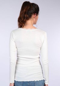 Weißes Langarmoberteil mit geripptem Textur, einer Spitzenborte am Ausschnitt und welligem Saum. Der Rücken hat eine taillierte Silhouette.