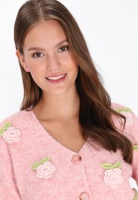 Rožinė megzta kardiganas su floristiniais aplikacijomis ir žaliais lapais, V formos iškirpte ir apvaliais mygtukais. Audinys turi minkštą, tekstūruotą paviršių.