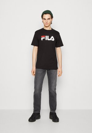 Zwarte t-shirt met rood en wit "FILA" logo, gecombineerd met grijze jeans en zwarte sneakers. Model staat tegen een effen achtergrond.
