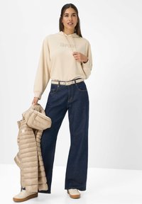 Beige sweatshirt met "Brox" logo, gecombineerd met losse donkerblauwe jeans. Houdt een beige pufferjack vast en draagt lichte sneakers met bruine zolen.
