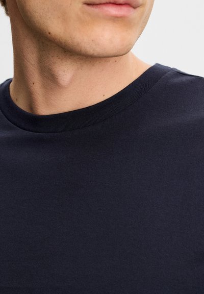 Κοντινό πλάνο του κάτω μέρους του προσώπου και του λαιμού ενός ατόμου που φοράει μονόχρωμη σκούρα μπλε μπλούζα με στρογγυλή λαιμόκοψη πάνω σε ανοιχτό φόντο.