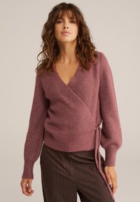Žena s valovitom smeškom kestenjastom kosom nosi mauve pleteni omotalac s dugim rukavima i smešnim hlačama s prugama na smešnoj pozadini.