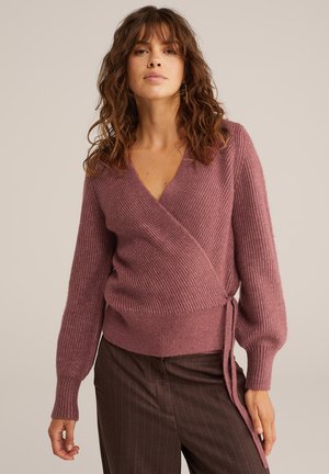 Femme aux cheveux bruns ondulés portant un pull mauve à côtes à manches longues et un pantalon marron à rayures contre un fond uni.