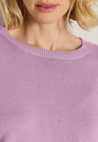 Frau mit kurzem blonden Haar, die einen helllila gestrickten Pullover mit rundem Ausschnitt trägt.