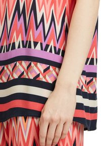 Haut à motifs multicolores avec des designs géométriques en corail, rose, marine et crème. Tissu lisse avec une coupe ample.