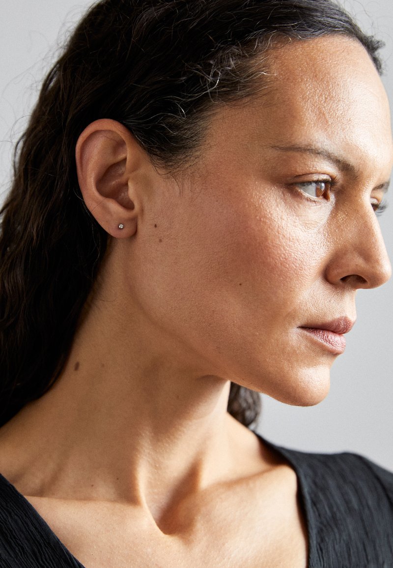 Mali, srebrni stakasti naušnica na lijevom uhu žene s srednjom smeškom kožom, dugom tamnom kosom i teksturiranim crnim topom.